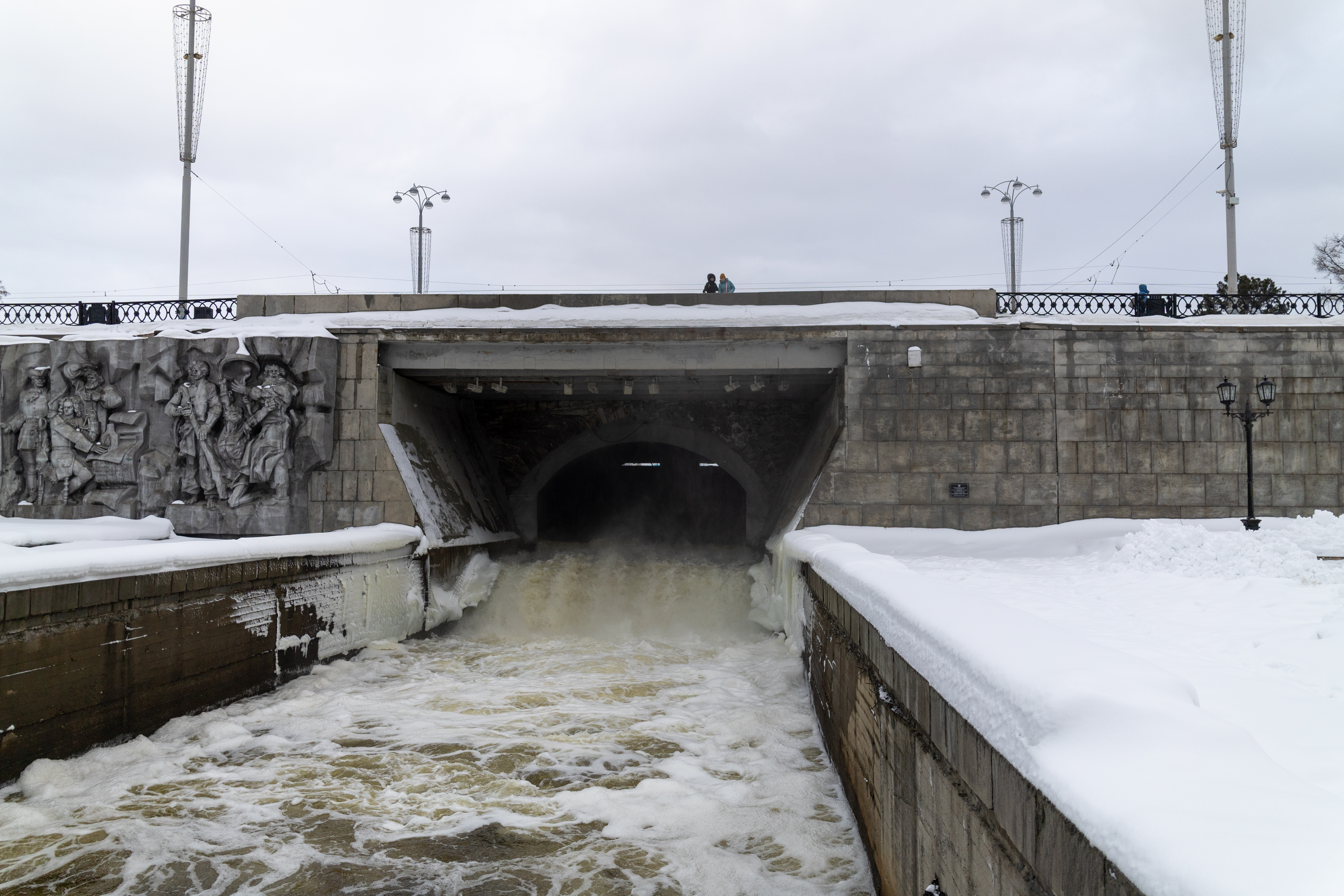 Уличный арт во дворе старых кирпичных зданий: мурал с изображением старика-ремесленника, яркие краски на фоне снега и красного кирпича.
