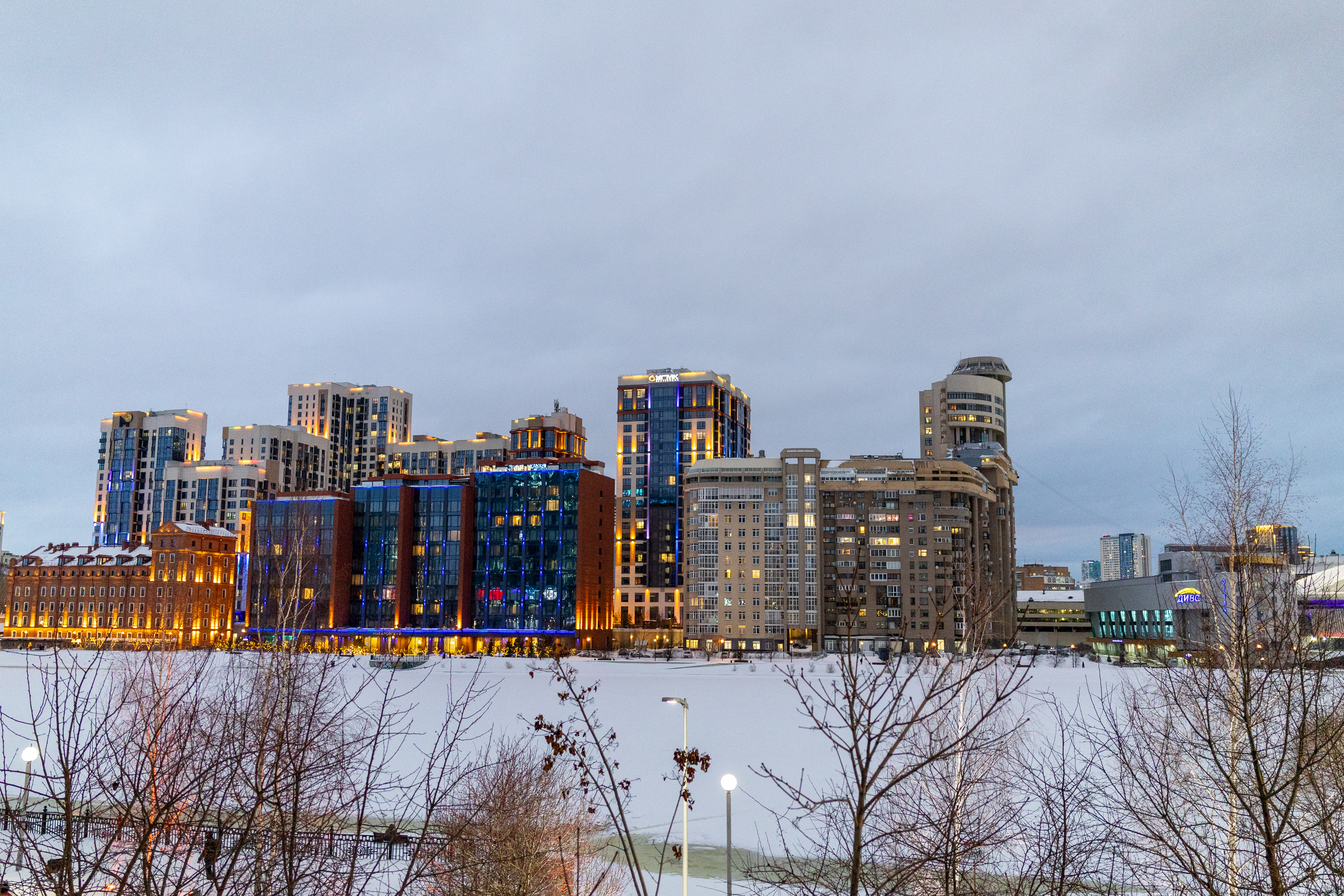 Ельцин Центр и набережная в сумерках: стеклянные башенки, подсветка арок, мост через Исеть с розоватой подсветкой, огни делового квартала.