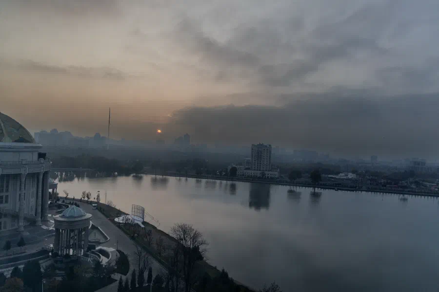 Закатное солнце пробивается сквозь облака над озером и силуэтом города